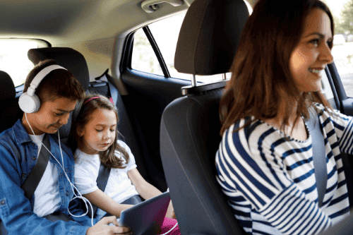 Get Ready for Your Travels with Our Road Trip Checklist with Little Wolf Automotive in Waupaca Wi, image of smiling mother driving car with son and daughter in backseat with headphones on and playing on a tablet during road trip