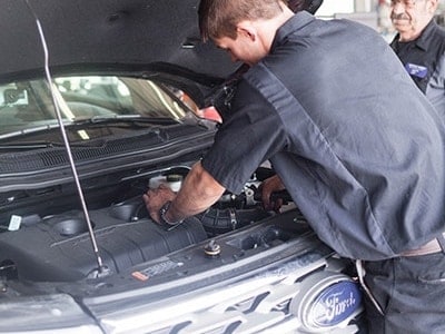 Engine service at Little Wolf Automotive image of back of mechanic holding hose of engine on ford vehicle in shop with hood open