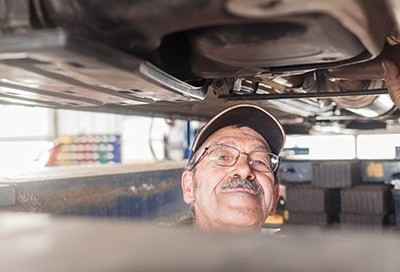 Vehicle inspection service | Little Wolf Automotive image of mechanic smiling while standing underneath car chassis that is raised on lift in shop