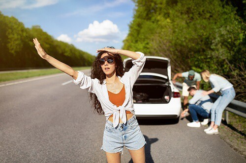 Locating an Express Auto Repair Station in Antigo, WI While on Vacation | Little Wolf Automotive in Antigo, WI. Image of a young woman trying to wave down cars, asking for help while her friends are trying to fix their broken vehicle while on the way to a vacation spot.