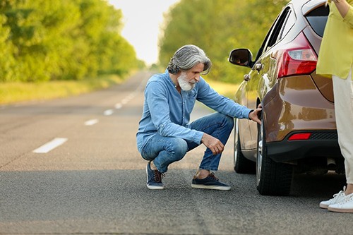 Mature man checking tires while on the road while female companion looks on. Concept image of “Avoid These 7 Common Pre-Trip Inspection Mistakes” | Little Wolf Auto in Waupaca, WI.