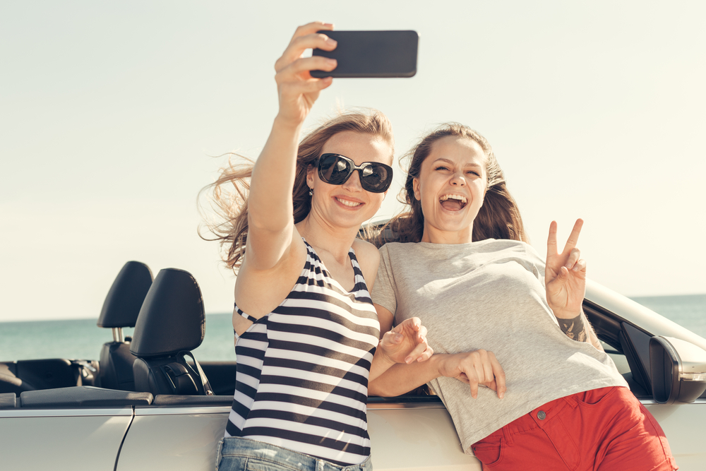 Summer Road Trip Essentials by Little Wolf Automotive Express Lube: Two friends taking a selfie by their car, symbolizing the joy and excitement of a well-prepared summer road trip, ensuring both safety and fun with proper car maintenance and essentials.